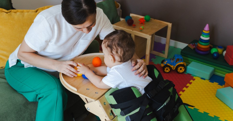 Terapeuta auxilia bebê posicionado em cadeira adaptada em atendimento domiciliar infantil.